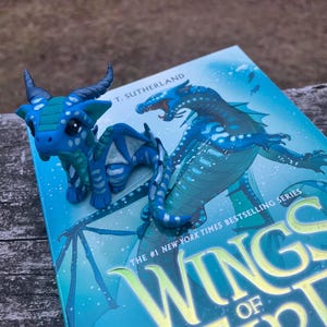 May include: A blue and teal dragon figurine rests on a book titled "Wings of Fire." The figurine has a detailed design with horns and wings. The book cover features a blue dragon illustration and the text "The #1 New York Times Bestselling Series."