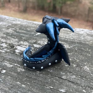 May include: A handcrafted, black sculpture of a creature with blue accents. The sculpture has a stylized design with white dot details. The creature appears to be resting on a wooden surface. The sculpture's design is unique and artistic.