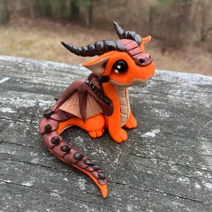 May include: A handcrafted orange and brown dragon figurine with large black eyes, horns, and wings. The dragon is made of clay and has a detailed, textured design. The figurine is sitting on a wooden surface.