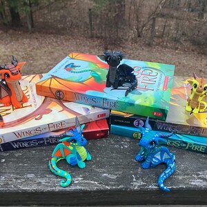 May include: A collection of colorful dragon figurines atop several books titled "Wings of Fire". The dragons are in various colors, including orange, blue, and yellow. The books have vibrant covers with dragon illustrations.