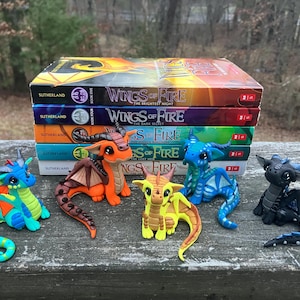 May include: A stack of "Wings of Fire" books with colorful dragon figurines. The figurines are in various colors, including blue, orange, yellow, and black. The books are stacked on a wooden surface.