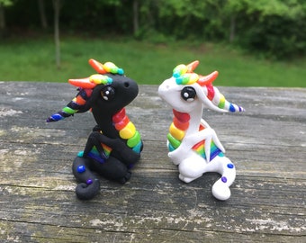 Op de afbeelding: Twee kleine, handgemaakte drakenbeeldjes. Eén is zwart met regenboogaccenten, de andere is wit met regenboogaccenten. Beide hebben grote, ronde ogen en staan op een houten oppervlak. De draken hebben regenbooghoorns en vleugels.