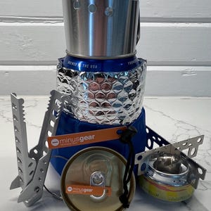 May include: A compact camping stove made from a blue can with a silver reflective wrap. It includes a silver windscreen, a small burner, and a lid. The stove has the text "minusgear" on a small orange tag. The can says "Brewed in the USA".