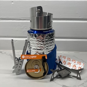 Könnte beinhalten: Ein kompaktes Campingkocher-Set mit einem silbernen Windschutz, einer blauen Isolierhülle und einer kleinen Brennstoffdose. Enthält einen silbernen Topfheber, einen kleinen Ständer und ein Brennstoffpäckchen. Das Set ist für das Kochen im Freien konzipiert.