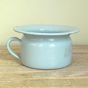 May include: A light blue enamel chamber pot featuring a wide, rounded rim and a single side handle. The vessel has a smooth, glossy finish with minor signs of wear along the edges, showcasing a classic, functional design for personal use.