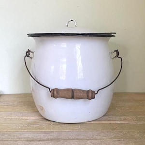May include: A white enamel pot with a black rim and lid. It has a wooden handle and a metal wire handle. The pot is sitting on a wooden surface.