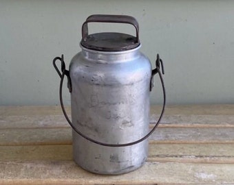 Puede incluir: Una antigua lata de leche de plata con asa de alambre. La lata tiene una tapa y está ligeramente abollada.