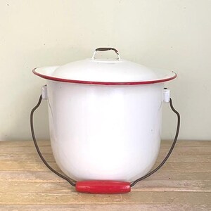 May include: A white enamel bucket with a matching lid, featuring a thin red rim around the top edge. The bucket has a dark metal swing handle with a bright red wooden grip, and a small metal handle on the center of the lid.