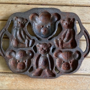 May include: A vintage cast iron bear-shaped muffin tin with six individual molds. The tin is rusty brown and has two handles.