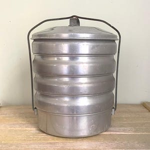 May include: A multi-tiered, cylindrical aluminum food carrier featuring three stacked compartments with a fitted lid and a sturdy, arched metal handle that pivots on the sides for easy transport and secure storage of food items.