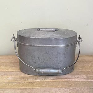 May include: An oval-shaped metal container with a weathered gray finish. It features a flat lid with a small handle and a larger, arched wire bail handle with a central wooden grip attached to the sides of the main body for carrying.