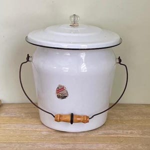 May include: White enamelware bucket with a lid and a handle. The bucket has a black rim and a wooden handle. A small sticker is on the front of the bucket. The lid has a clear, decorative knob. The bucket is likely vintage.