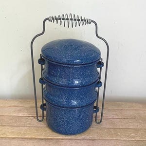 May include: A three-tiered stackable food container with a dark blue speckled enamel finish. The set features a metal frame with a coiled wire handle that secures the three round compartments and a matching lid in a vertical, portable arrangement.
