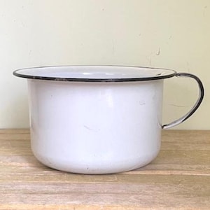 May include: A white enamelware bowl with a black rim and handle. The bowl is round with a slightly flared rim and a curved handle. The bowl is sitting on a wooden surface.
