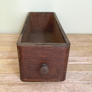 May include: A rectangular wooden drawer with a dark brown finish and a visible wood grain texture. The drawer features a single small, centered wooden knob on the front panel and an open top, revealing the hollow interior compartment.