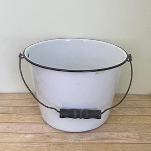 May include: A white enamel bucket with a black rim and handle. The bucket has a dark wooden handle and a metal wire handle. The bucket is sitting on a wooden surface.
