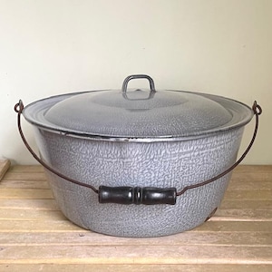 May include: A gray enamelware pot with a lid and a handle. The pot has a speckled pattern and a dark brown handle. The lid has a small handle. The pot is sitting on a wooden surface.