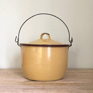 May include: A vintage yellow enamelware pot with a lid and a black wire handle. The pot has a rounded shape with a dark brown trim around the rim and lid. The handle arches over the top, attached to the sides.