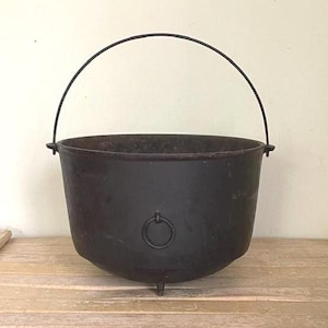 May include: A black cast iron cauldron with a curved handle. The cauldron has a rounded base, three small feet, and a ring detail on the front. The handle is made of a dark metal. The cauldron is sitting on a wooden surface.
