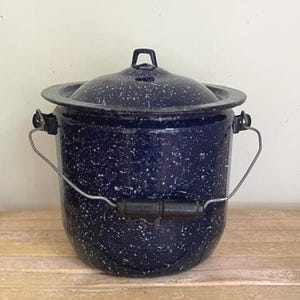 May include: A dark blue enamel pot with a white speckled pattern, featuring a matching lid with a small handle, and a metal bail handle with a black wooden grip. The cylindrical container has a wide rim and a sturdy construction for storage.