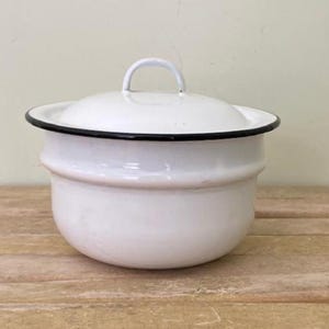 May include: A white enamelware pot with a lid. The pot has a black rim and a white handle on the lid. The pot is sitting on a wooden surface.