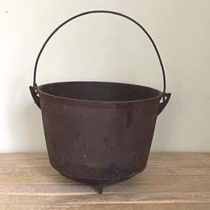 May include: A dark brown cast iron cauldron with a curved handle. The cauldron has three small legs and is sitting on a wooden surface. The handle is made of a dark metal. The cauldron is likely used for cooking or decorative purposes.