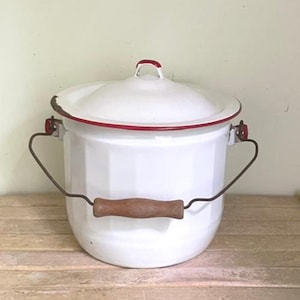 May include: A white enamel bucket with a matching lid, featuring a thin red rim and a red handle accent on the lid. The bucket has a metal wire bail handle with a central wooden grip and shows signs of wear along the upper rim edge.