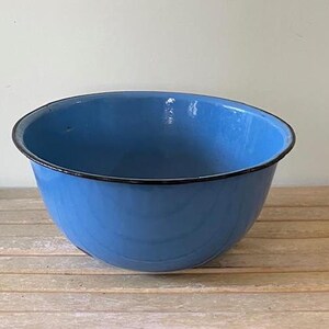 May include: A large, light blue enamelware bowl with a black rim. The bowl is sitting on a wooden surface. The bowl is a vintage kitchenware item.