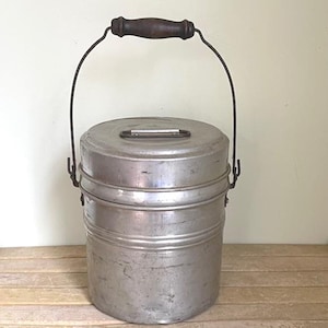May include: Vintage metal bucket with a wooden handle. The bucket is cylindrical with a lid and a wire handle. The metal has a worn, aged appearance. The handle is dark brown.