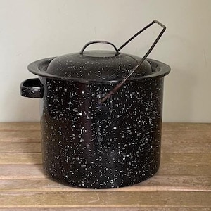 May include: A black enamel pot with a lid and a handle. The pot has a speckled white pattern and a metal handle that arches over the top. The pot sits on a wooden surface.