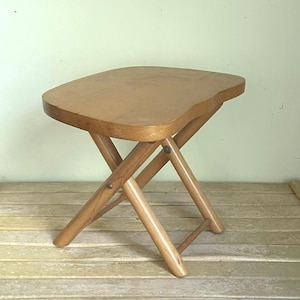 May include: A light brown, folding wooden stool with a curved seat. The legs are angled and cross in an X-shape, connected by metal rivets. The stool is approximately 15 inches tall.