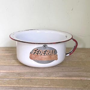 May include: Vintage white enamelware chamber pot with a red rim and handle. The pot features the text "FEDERAL VOGUE ENAME" on the front. The pot is sitting on a wooden surface.