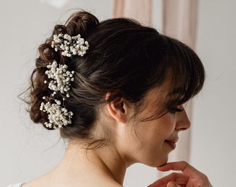 Könnte beinhalten: Eine Frau mit langen braunen Haaren, die zu einem Dutt mit weißen Babyschnabelblumen gesteckt sind. Sie trägt ein weißes Spitzenkleid mit tiefem Rücken.