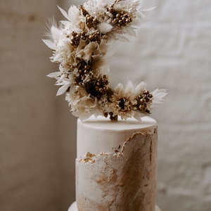 Op de afbeelding: Een tweelaagse bruidstaart met een witte en gouden afwerking. De bovenste laag heeft een droogbloemenarrangement in de vorm van een halve maan.