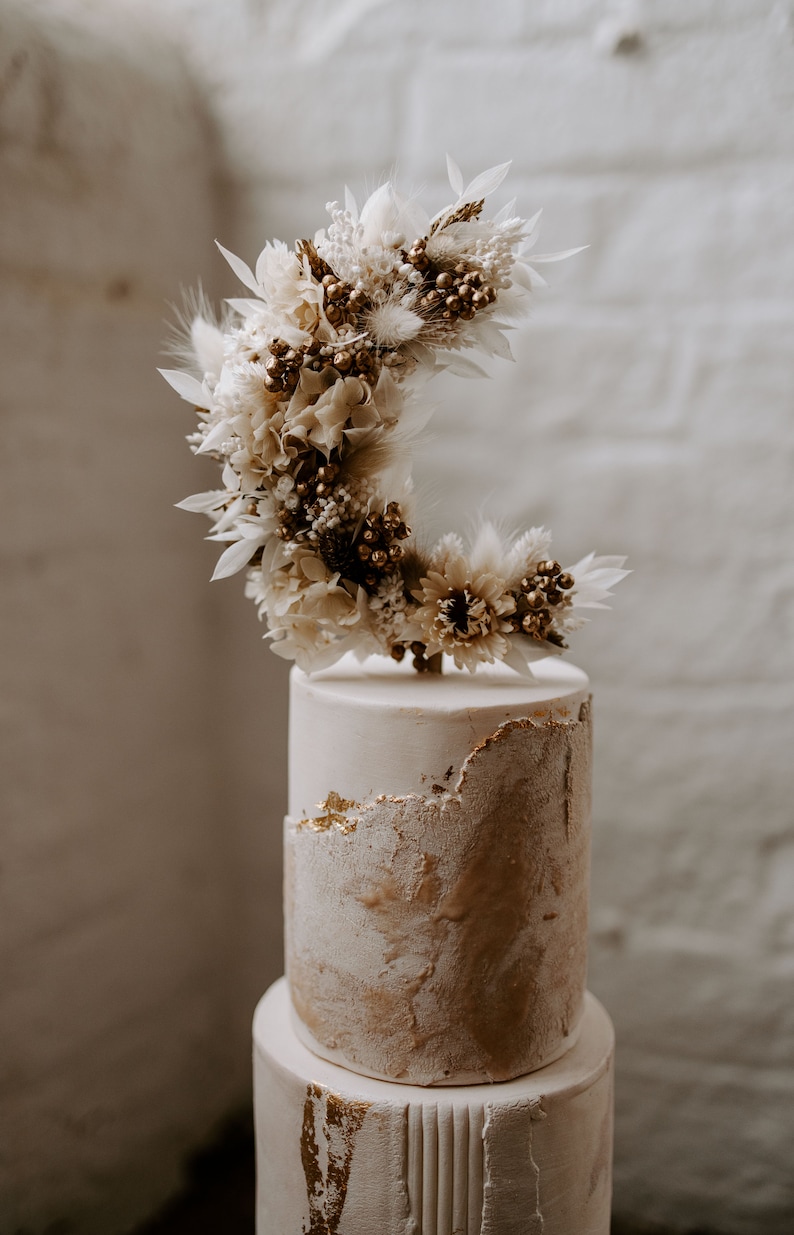 Op de afbeelding: Een tweelaagse bruidstaart met een witte en gouden afwerking. De bovenste laag heeft een droogbloemenarrangement in de vorm van een halve maan.