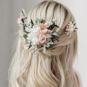 Op de afbeelding: Een bloemenhaar kam met witte en roze bloemen, groen en gedroogde grassen. De kam wordt gedragen in het blonde haar van een vrouw.