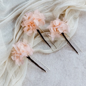 Op de afbeelding: Drie roze bloemen haarclips met zilveren en bruine pinnen. De clips liggen op een witte stoffen achtergrond.