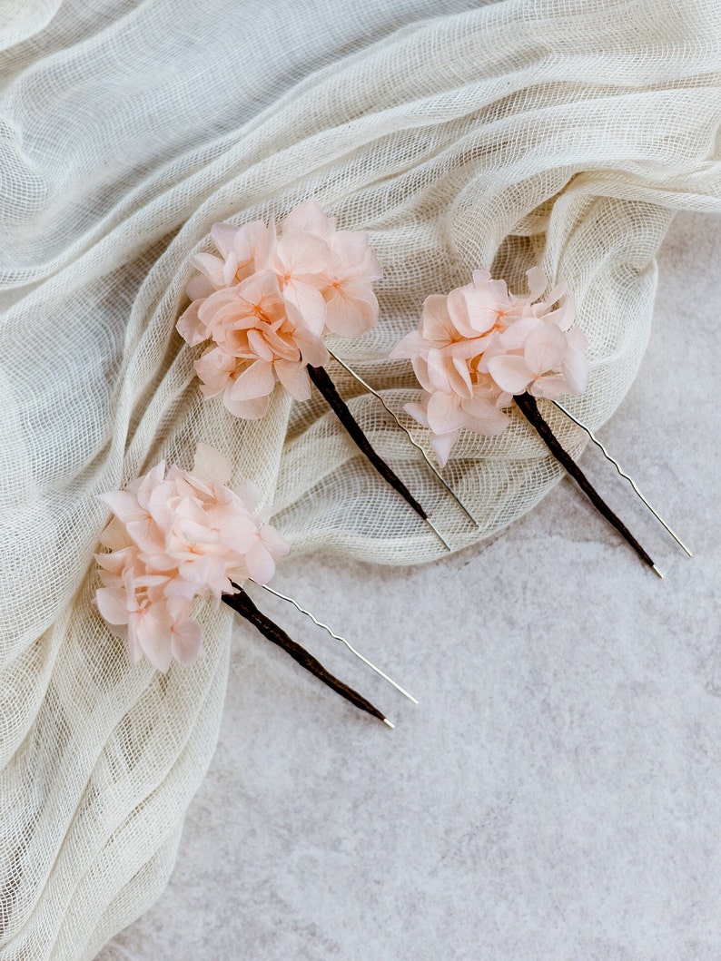 Op de afbeelding: Drie roze bloemen haarclips met zilveren en bruine pinnen. De clips liggen op een witte stoffen achtergrond.