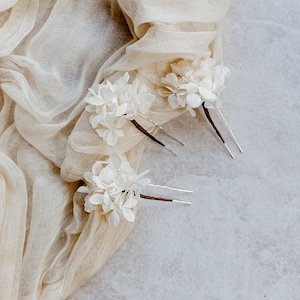 Op de afbeelding: Drie witte bloemenhaarspeldjes met zilveren pinnen. De bloemen zijn gemaakt van delicaat wit stof en zijn in een tros gerangschikt. De speldjes liggen op een beige stoffen achtergrond.