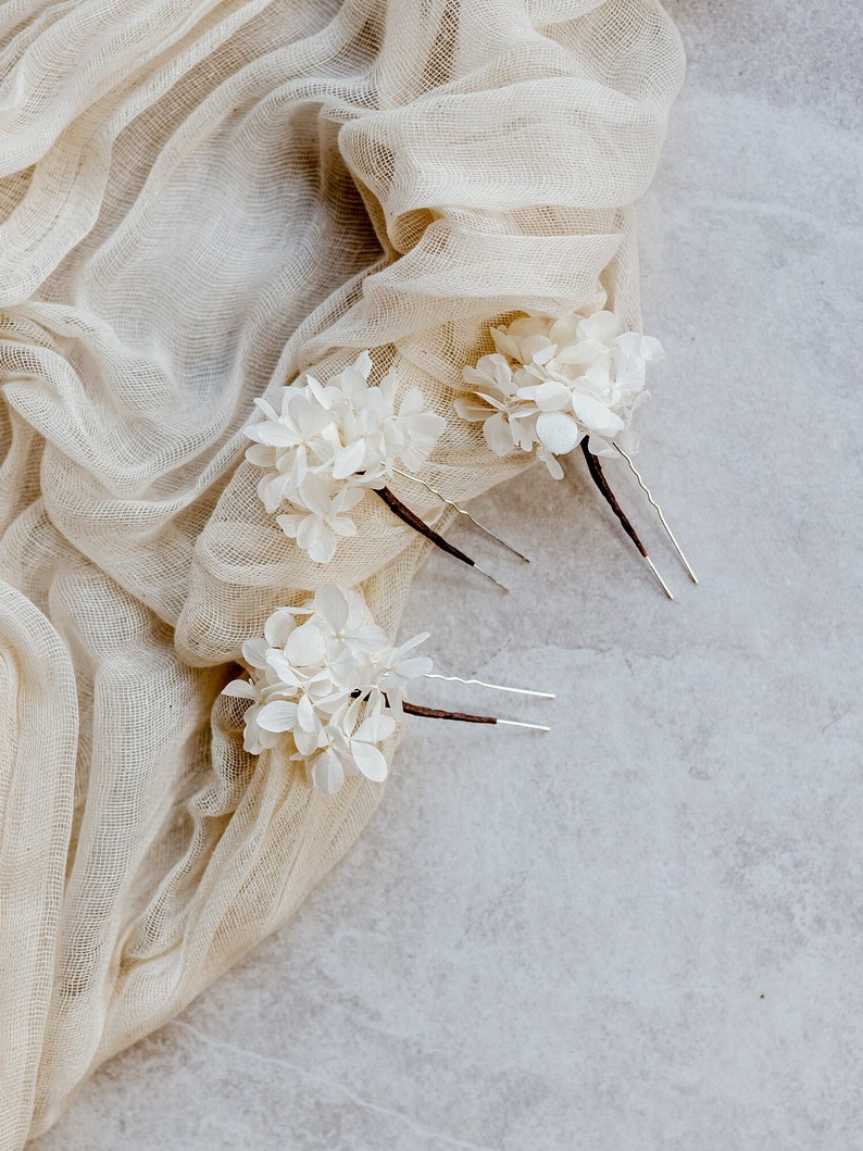 Op de afbeelding: Drie witte bloemenhaarspeldjes met zilveren pinnen. De bloemen zijn gemaakt van delicaat wit stof en zijn in een tros gerangschikt. De speldjes liggen op een beige stoffen achtergrond.