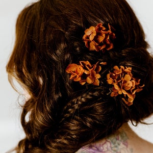 Op de afbeelding: Een vrouw met lang bruin haar dat in een vlecht is gevlochten met drie kleine bosjes gedroogde oranje bloemen.