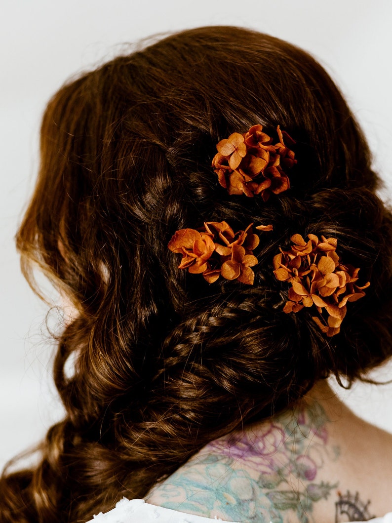 Op de afbeelding: Een vrouw met lang bruin haar dat in een vlecht is gevlochten met drie kleine bosjes gedroogde oranje bloemen.