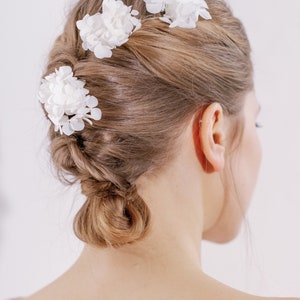 Op de afbeelding: Drie witte bloemen haarclips in het haar van een vrouw. Het haar is gestyled in een knot met een vlecht.