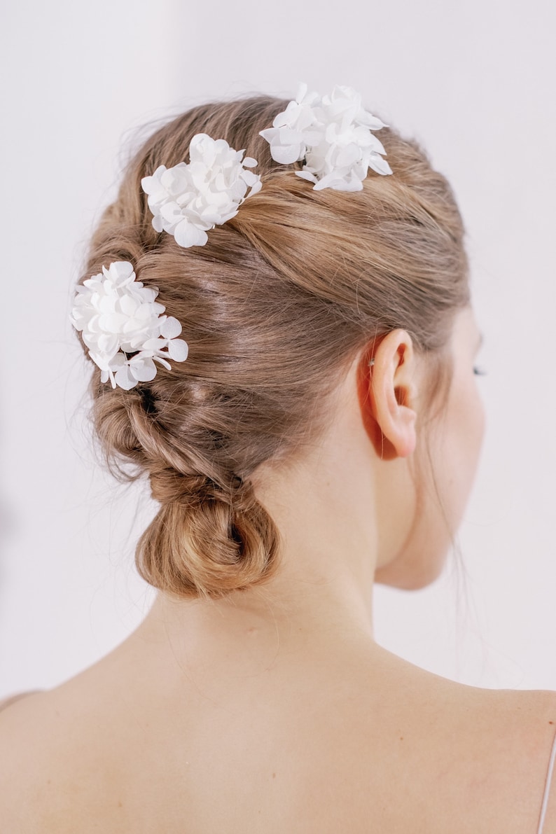 Op de afbeelding: Drie witte bloemen haarclips in het haar van een vrouw. Het haar is gestyled in een knot met een vlecht.