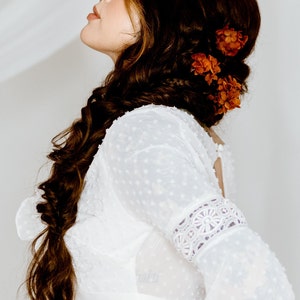 Op de afbeelding: Een vrouw met lang bruin haar dat in een vlecht is gevlochten met oranje bloemen erin geweven. Ze draagt een wit, met kant afgewerkt, getailleerd topje met een gestippeld patroon.