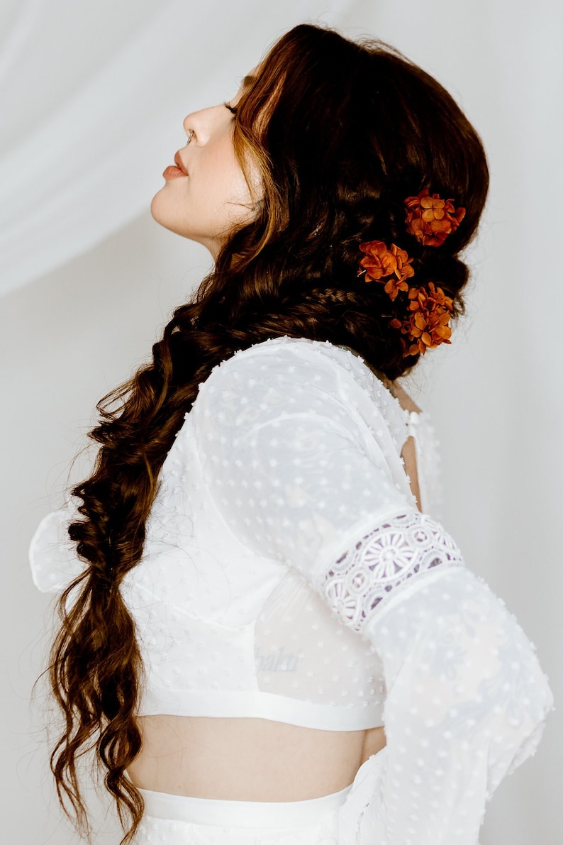 Op de afbeelding: Een vrouw met lang bruin haar dat in een vlecht is gevlochten met oranje bloemen erin geweven. Ze draagt een wit, met kant afgewerkt, getailleerd topje met een gestippeld patroon.