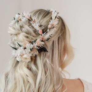 Op de afbeelding: Een bloemenkrans gemaakt van gedroogde bloemen in tinten wit, roze en bruin. De krans wordt gedragen op het hoofd van een persoon met blond haar.