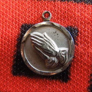 May include: A silver-toned pendant featuring a detailed depiction of praying hands. The round pendant has a textured border and a small loop for attachment. The background is a red and black patterned fabric.