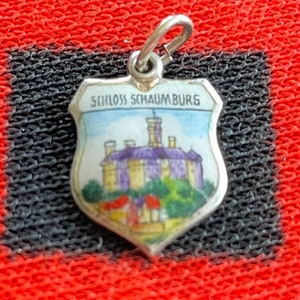 May include: A silver charm in the shape of a shield with a painted image of a castle and the text "Schloss Schaumburg" on it.