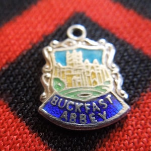 May include: Silver charm with blue enamel depicting Buckfast Abbey, a historic Benedictine monastery in Devon, England. The charm features a detailed illustration of the abbey building with a green lawn and a blue sky.